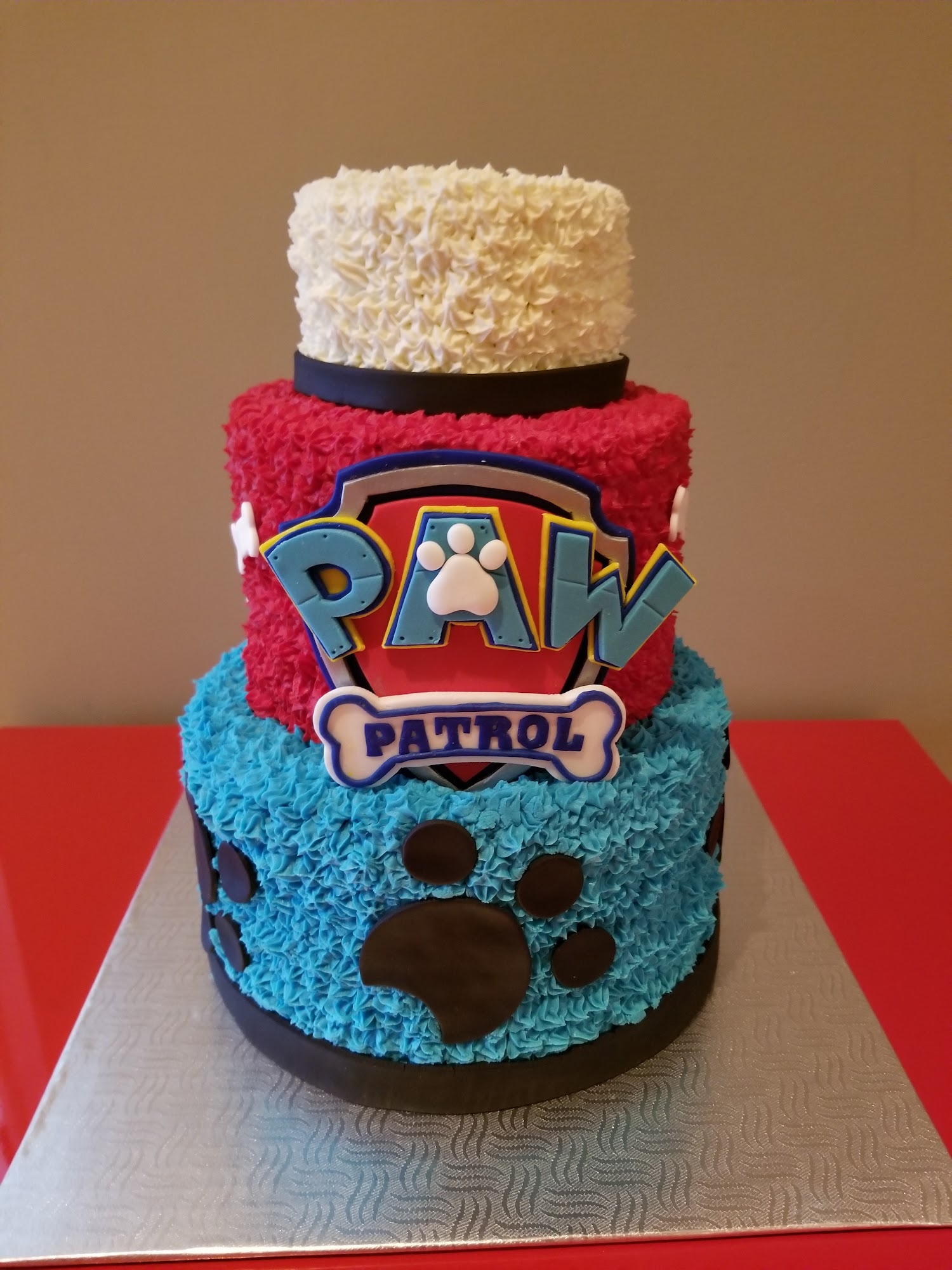 Gâteau trois étages Pat' Patrouille, glaçage froufrouté blanc, rouge et bleu, logo au centre et empreintes de pattes.